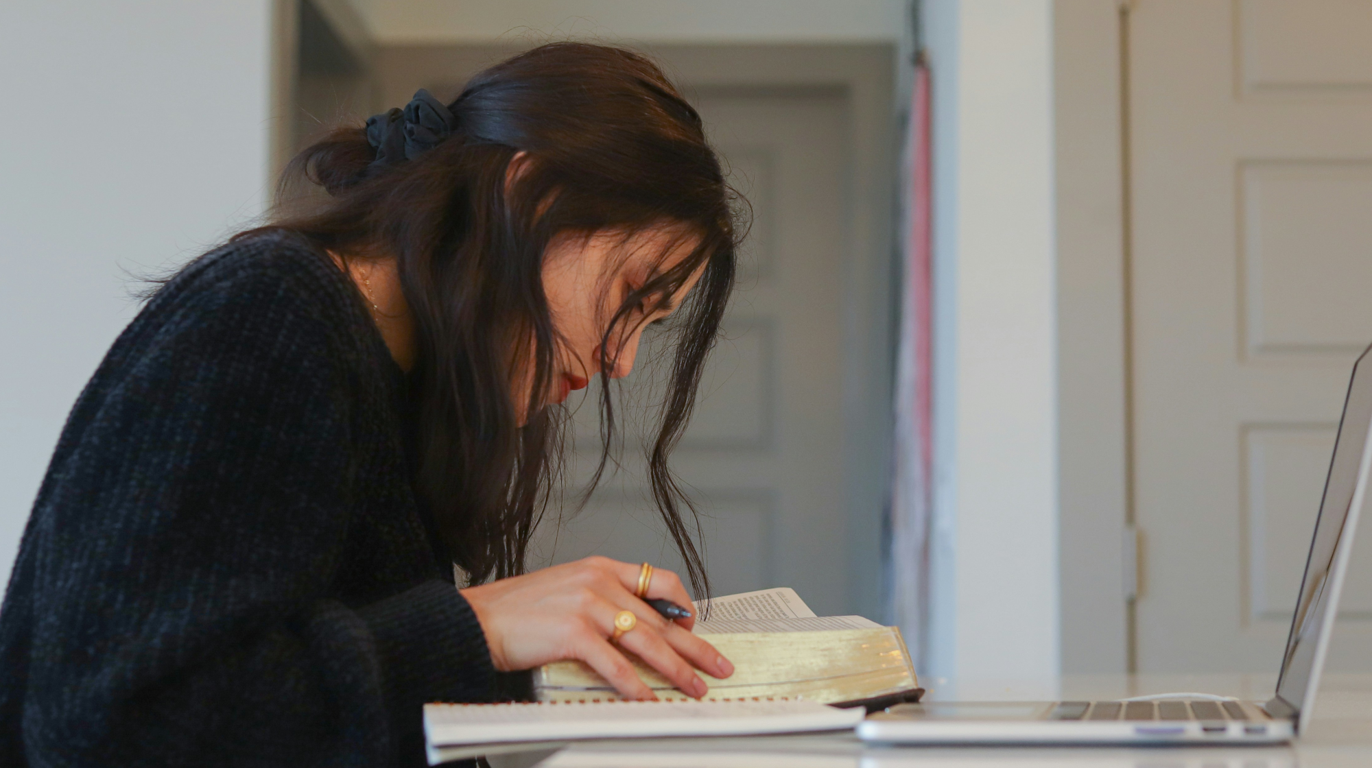Woman Looking at a Book with a Pen, Notebook, and Laptop